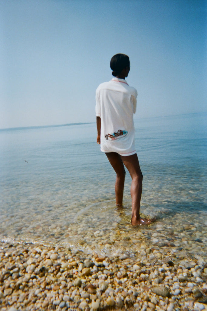 Woman standing in shallow water with her back to us, wearing the white terry short-sleeve 'Boston Whaler' Cabana, which features a boat illustration on the back.