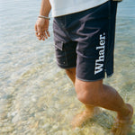 A man stands ankle-deep in the ocean, wearing navy board shorts with white contrast stitching and white 'Whaler' text on the left leg.