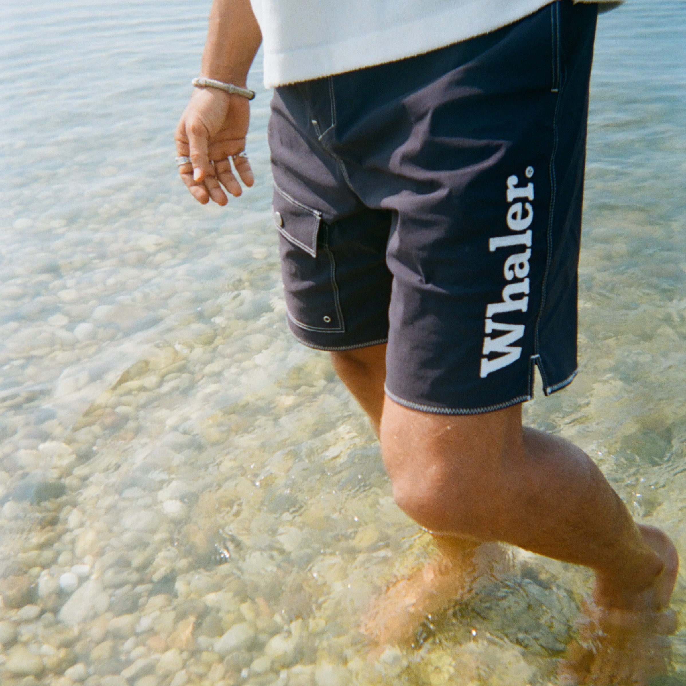 A man stands ankle-deep in the ocean, wearing navy board shorts with white contrast stitching and white 'Whaler' text on the left leg.