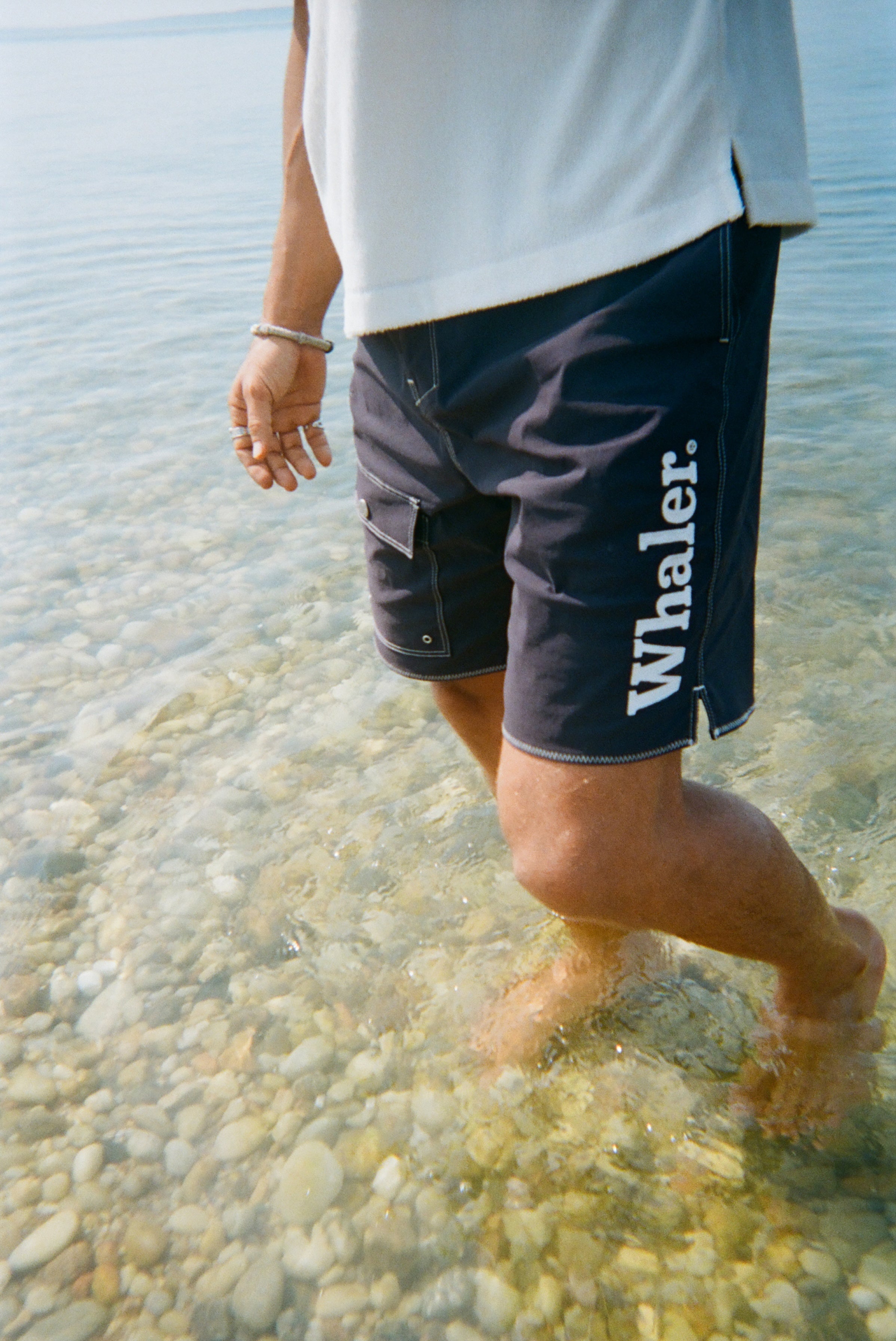 A man stands ankle-deep in the ocean, wearing navy board shorts with white contrast stitching and white 'Whaler' text on the left leg.