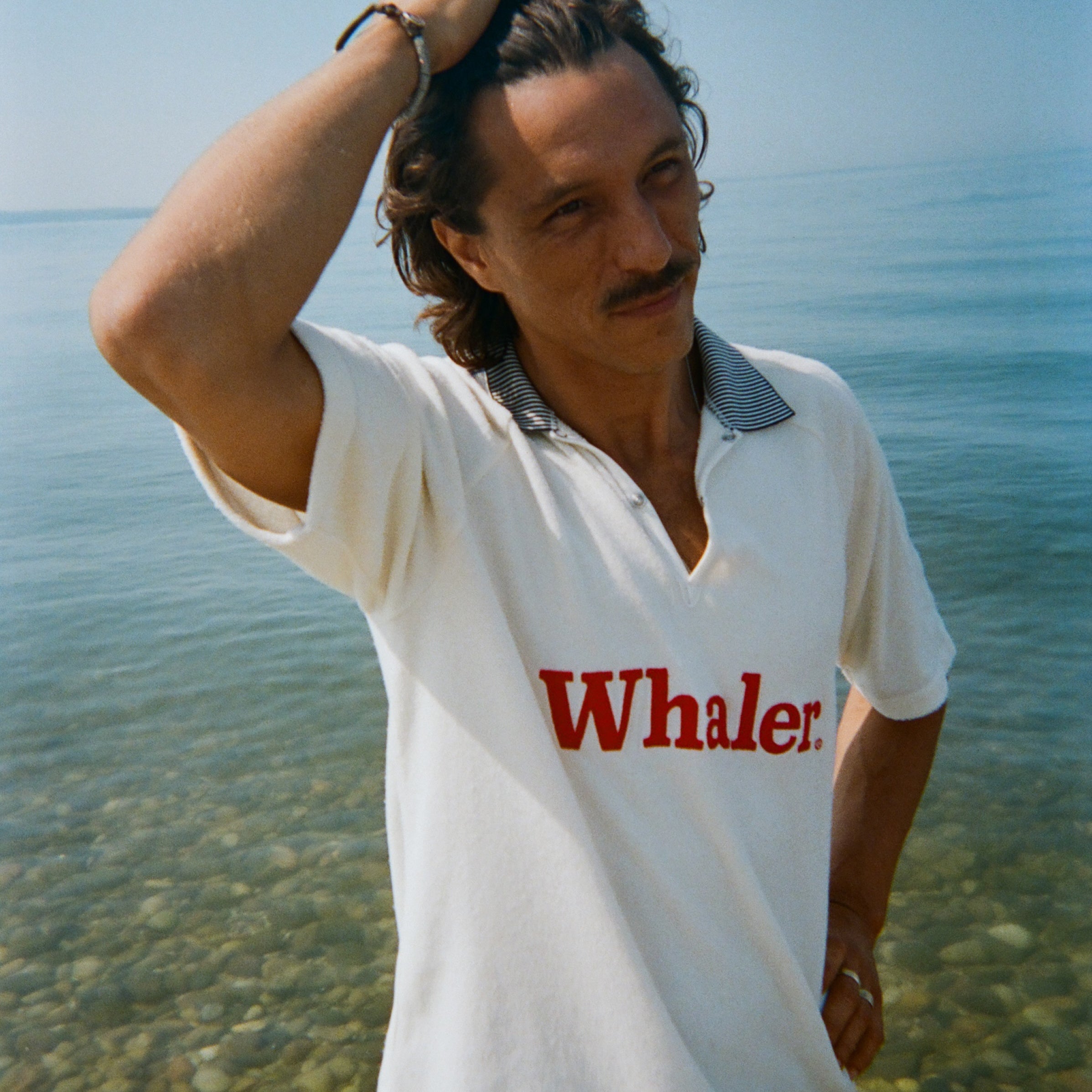 A brunette man stands in shallow water, wearing the white short-sleeve terry polo with red 'Whaler' text across the chest and a black-and-white striped collar.