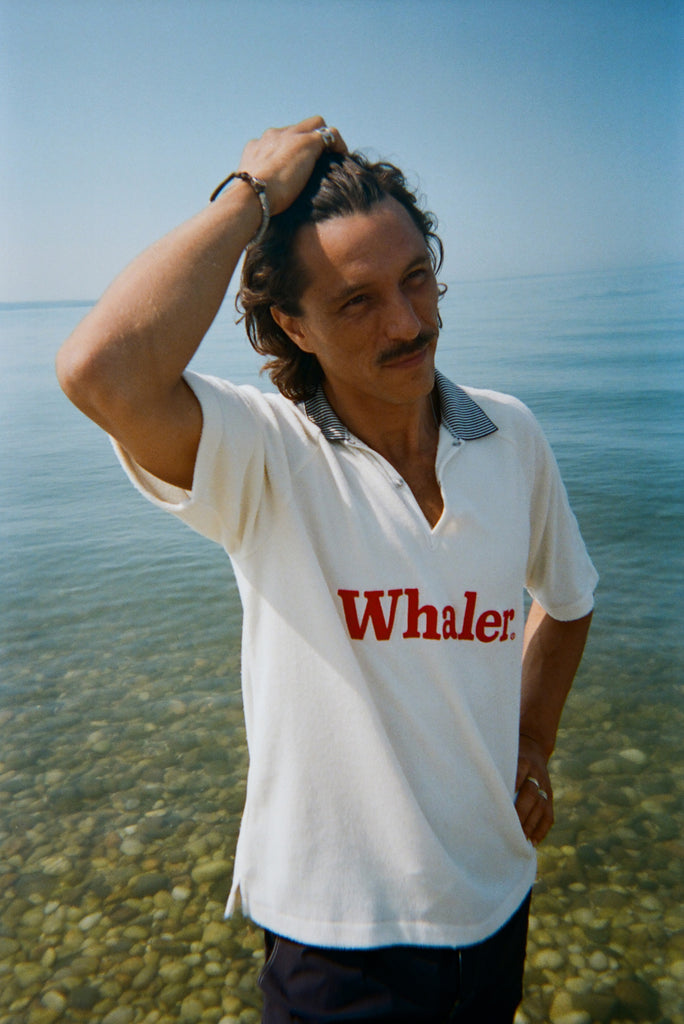 A brunette man stands in shallow water, wearing the white short-sleeve terry polo with red 'Whaler' text across the chest and a black-and-white striped collar.
