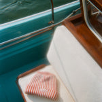 Red and white striped knit beanie with a folded brim, featuring a 'Whaler The Unsinkable Legend' patch on the front, resting on a cream boat cushion.