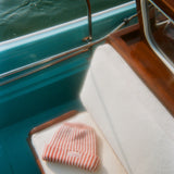 Red and white striped knit beanie with a folded brim, featuring a 'Whaler The Unsinkable Legend' patch on the front, resting on a cream boat cushion.