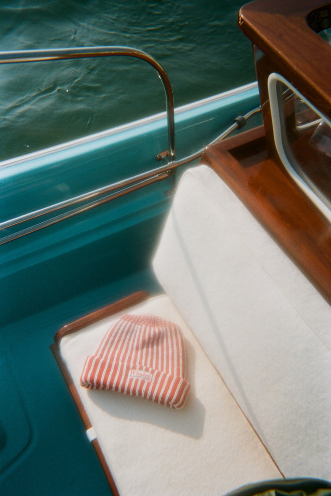 Red and white striped knit beanie with a folded brim, featuring a 'Whaler The Unsinkable Legend' patch on the front, resting on a cream boat cushion.