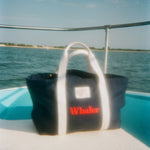 Navy canvas tote bag with white handles and a front pocket is seen sitting on a boat. Red embroidered text on the pocket reads ‘Whaler.’ A small white label above the pocket reads ‘Wm. J. Mills & Co. Sailmakers, Greenport, N.Y.’