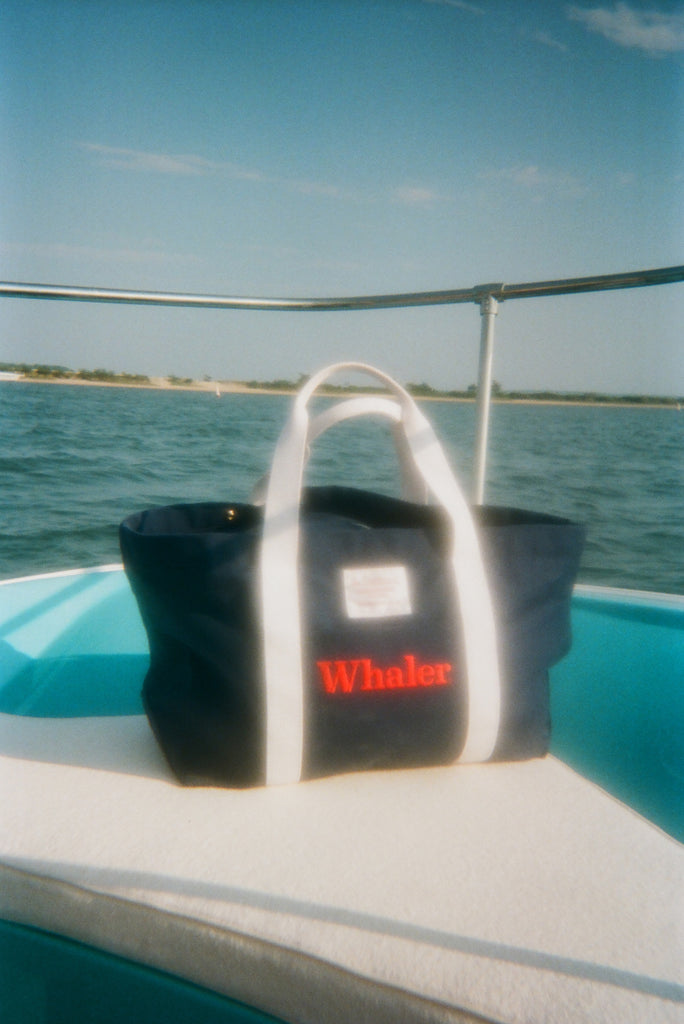 Navy canvas tote bag with white handles and a front pocket is seen sitting on a boat. Red embroidered text on the pocket reads ‘Whaler.’ A small white label above the pocket reads ‘Wm. J. Mills & Co. Sailmakers, Greenport, N.Y.’