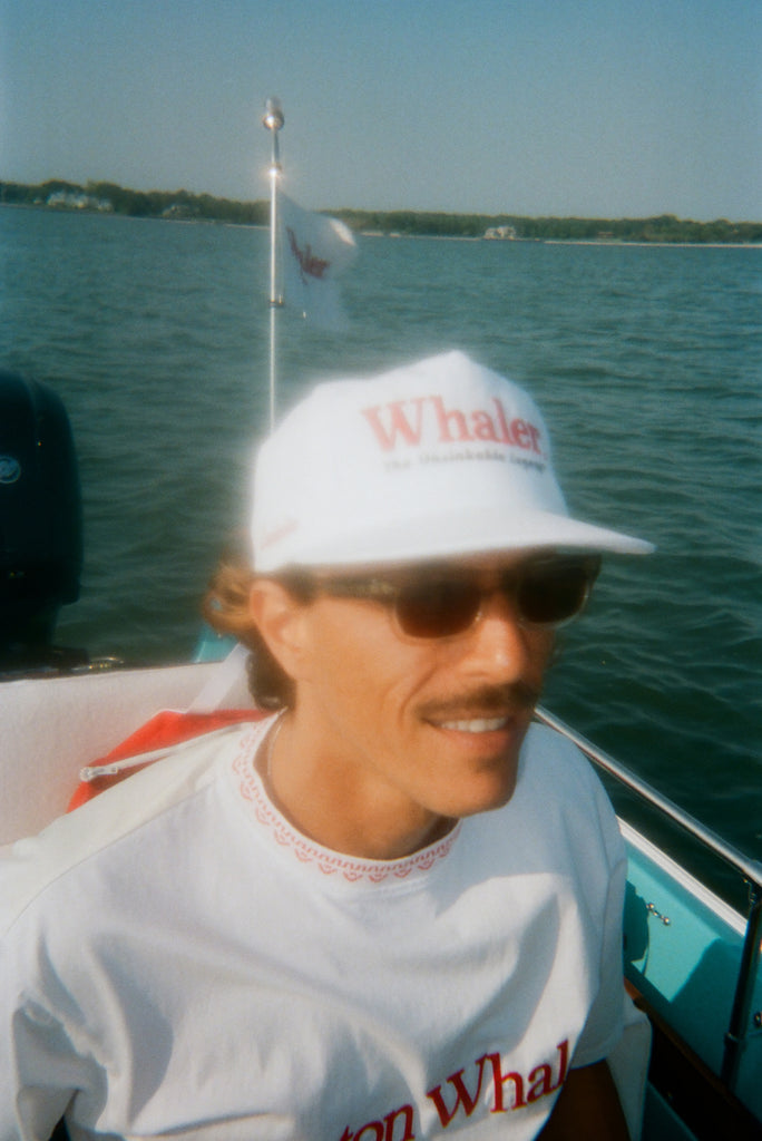 Close-up of a man sitting on a boat, wearing a white five-panel cap with red embroidered text reading 'Whaler' and smaller text below reading 'The Unsinkable Legend'. He also wears dark sunglasses and a white 'Boston Whaler' shirt with red neckline detailing. 'Whaler' flag is visible in the background.