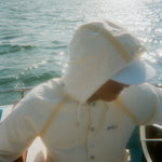 A man is pictured sitting on a boat wearing a hooded cream jacket with tan trim and white board shorts. A white cap is visible under the hood. 