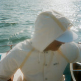 A man is pictured sitting on a boat wearing a hooded cream jacket with tan trim and white board shorts. A white cap is visible under the hood. 