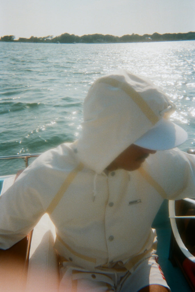 A man is pictured sitting on a boat wearing a hooded cream jacket with tan trim and white board shorts. A white cap is visible under the hood. 
