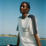 Woman sitting on a boat wearing a light blue cotton mesh shirt with navy shoulder panels, a striped ribbed collar, and a navy chest pocket with white 'Boston Whaler' embroidery. She pairs it with navy shorts with white trim.