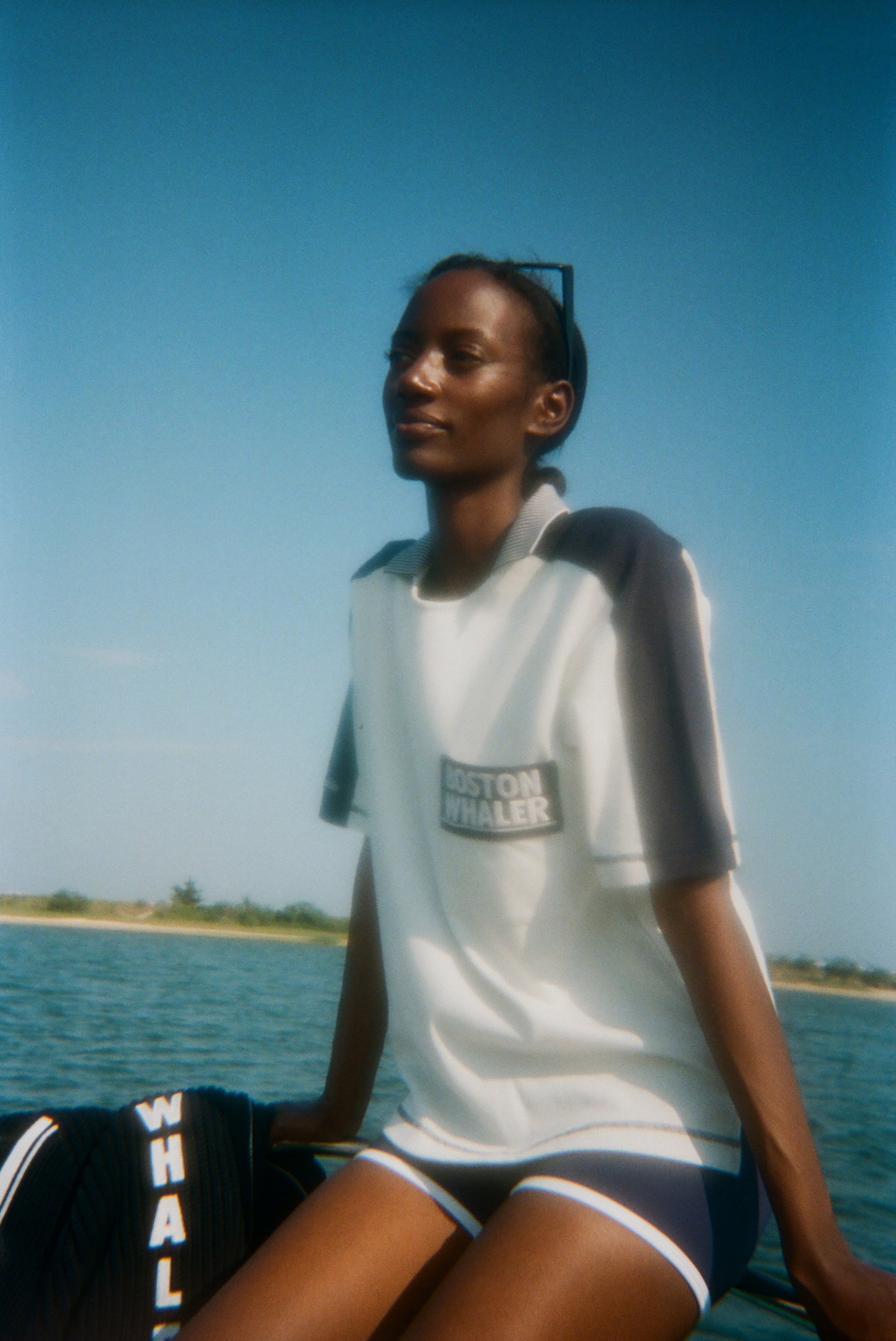 Woman sitting on a boat wearing a light blue cotton mesh shirt with navy shoulder panels, a striped ribbed collar, and a navy chest pocket with white 'Boston Whaler' embroidery. She pairs it with navy shorts with white trim.