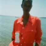 Woman standing at the water’s edge wearing a red button-up shirt with white contrast stitching and a mesh zip pocket on the chest. Sunglasses are perched on her head.