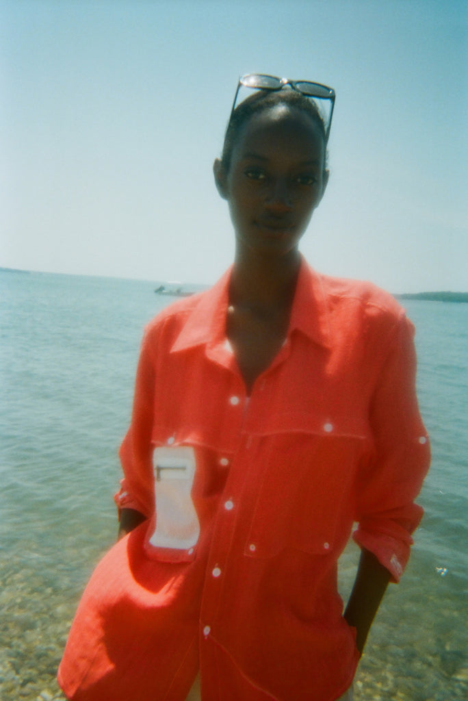 Woman standing at the water’s edge wearing a red button-up shirt with white contrast stitching and a mesh zip pocket on the chest. Sunglasses are perched on her head.