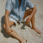 Man sitting on the sand in a light blue short-sleeve zip-up shirt with red and green button details. Shirt is partially unzipped to reveal his chest. 