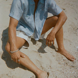Man sitting on the sand in a light blue short-sleeve zip-up shirt with red and green button details. Shirt is partially unzipped to reveal his chest. 