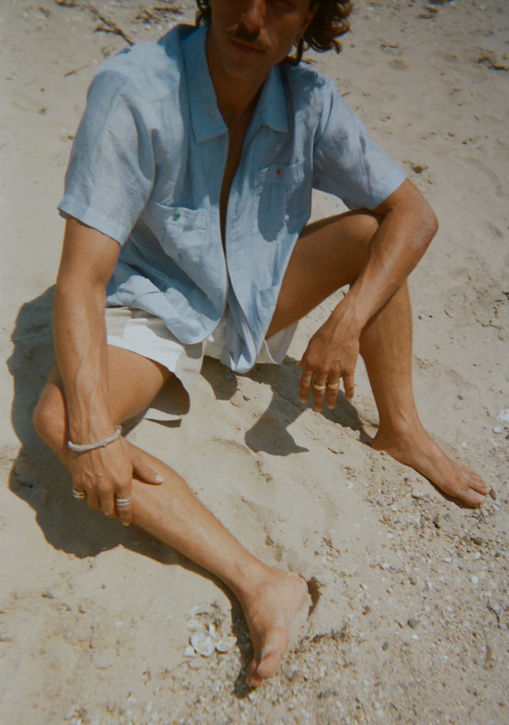 Man sitting on the sand in a light blue short-sleeve zip-up shirt with red and green button details. Shirt is partially unzipped to reveal his chest. 