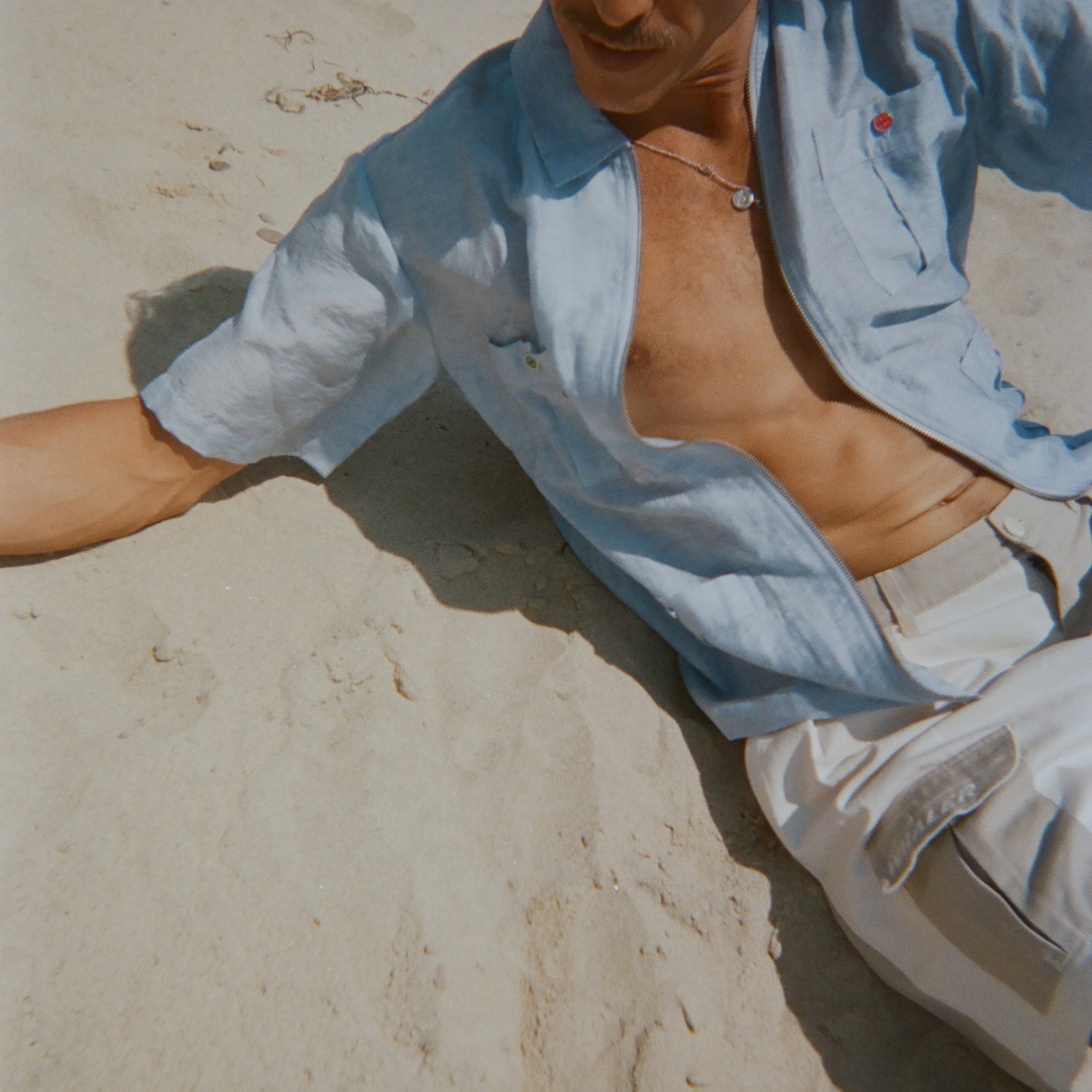 Man lounging on the sand in a light blue short-sleeve zip-up shirt with red and green button details. Shirt is partially unzipped to reveal chest and necklace.