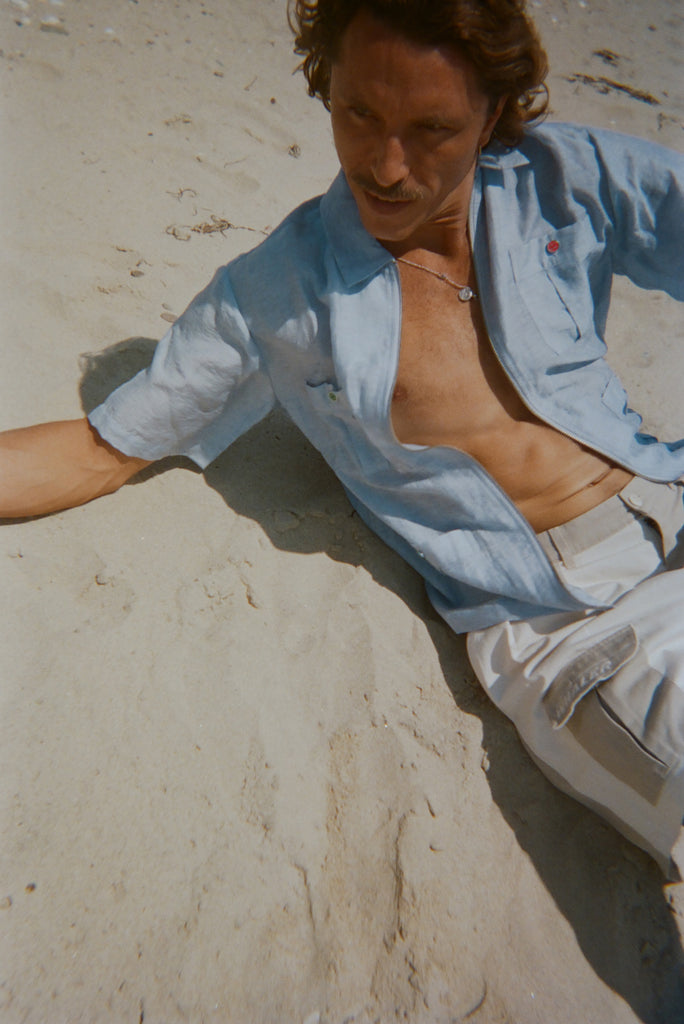 Man lounging on the sand in a light blue short-sleeve zip-up shirt with red and green button details. Shirt is partially unzipped to reveal chest and necklace.