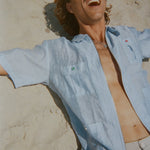 Man lounging on the sand in a light blue short-sleeve zip-up shirt with red and green button details. Shirt is partially unzipped to reveal chest and necklace.