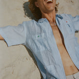 Man lounging on the sand in a light blue short-sleeve zip-up shirt with red and green button details. Shirt is partially unzipped to reveal chest and necklace.