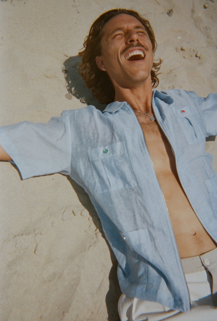 Man lounging on the sand in a light blue short-sleeve zip-up shirt with red and green button details. Shirt is partially unzipped to reveal chest and necklace.