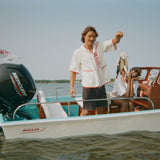 Man wearing a white hooded beach towel top with red text reading 'Whaler' holds up fish while standing on a Whaler boat. The woman sits beside him at the helm, smiling and leaning her chin on her hands.