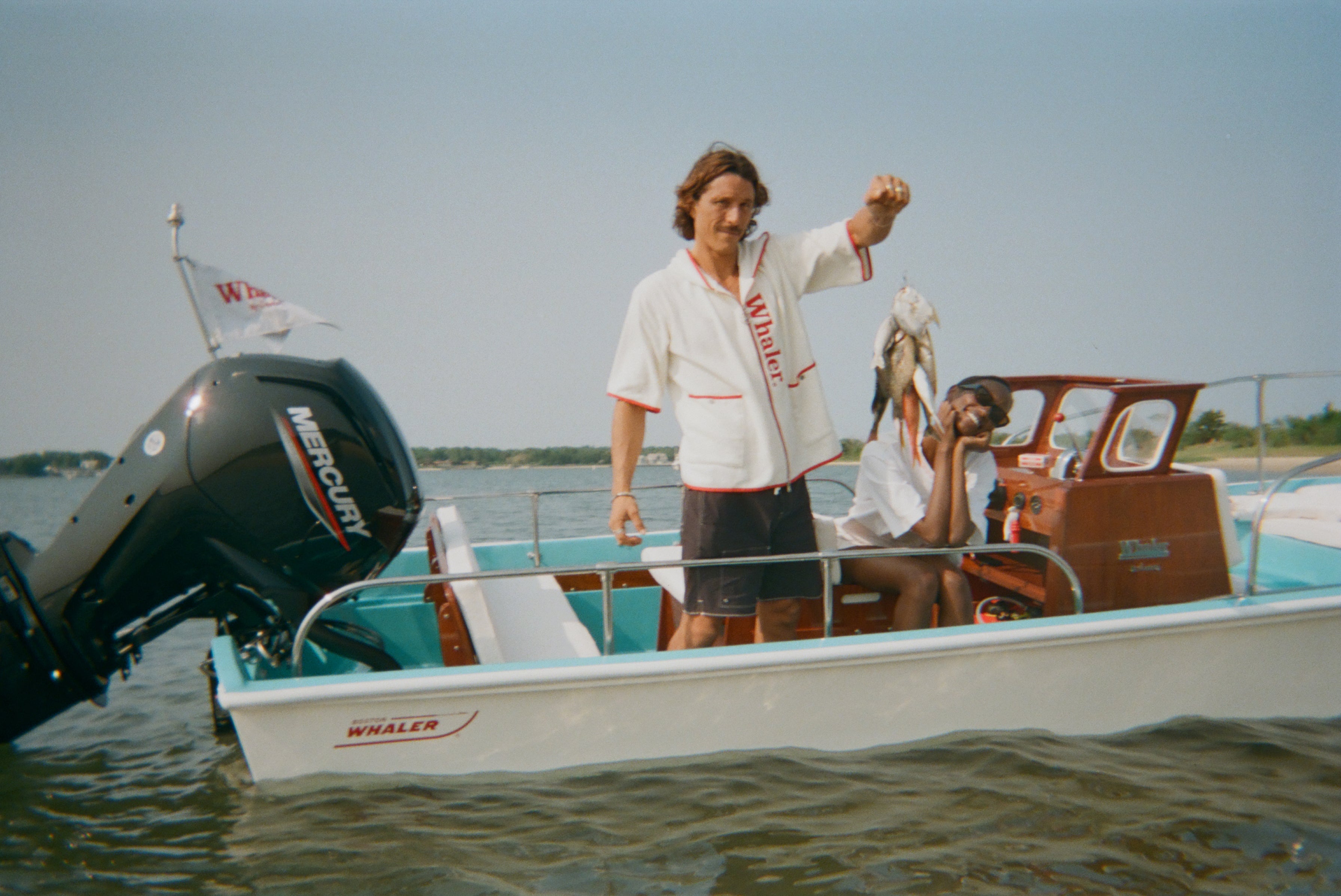 Man wearing a white hooded beach towel top with red text reading 'Whaler' holds up fish while standing on a Whaler boat. The woman sits beside him at the helm, smiling and leaning her chin on her hands.