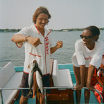 Man smiling while holding up three fish on a line, wearing a white zip-up shirt with red trim and vertical 'Whaler' text. Woman beside him leans on the boat's edge, wearing a white t-shirt with 'Boston Whaler' in red and a red knit collar.