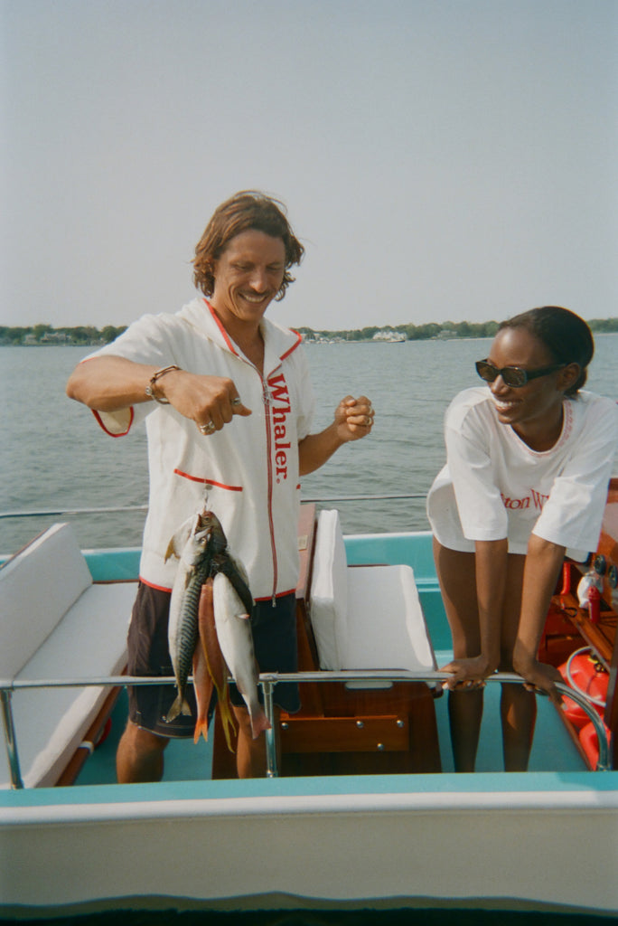 Man smiling while holding up three fish on a line, wearing a white zip-up shirt with red trim and vertical 'Whaler' text. Woman beside him leans on the boat's edge, wearing a white t-shirt with 'Boston Whaler' in red and a red knit collar.