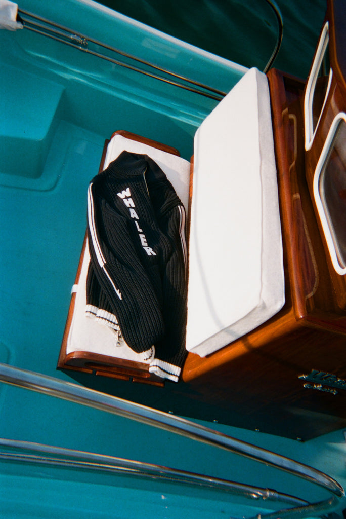 A navy ribbed cardigan with white 'WHALER' lettering is draped over a cushioned boat seat.