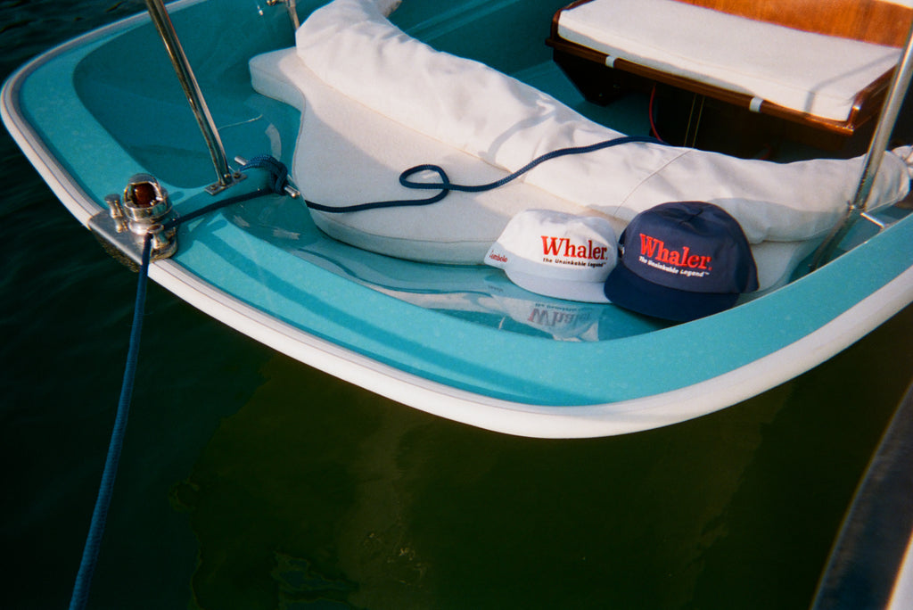 White and blue five-panel 'Whaler' caps with embroidered text reading 'The Unsinkable Legend' rest at the front of a light blue boat tied with a rope.