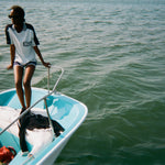 Woman standing at the edge of a boat wearing a light blue cotton mesh shirt with navy shoulder panels and a 'Boston Whaler' logo on the chest, paired with matching navy shorts with white trim. 