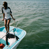 Woman standing at the edge of a boat wearing a light blue cotton mesh shirt with navy shoulder panels and a 'Boston Whaler' logo on the chest, paired with matching navy shorts with white trim. 