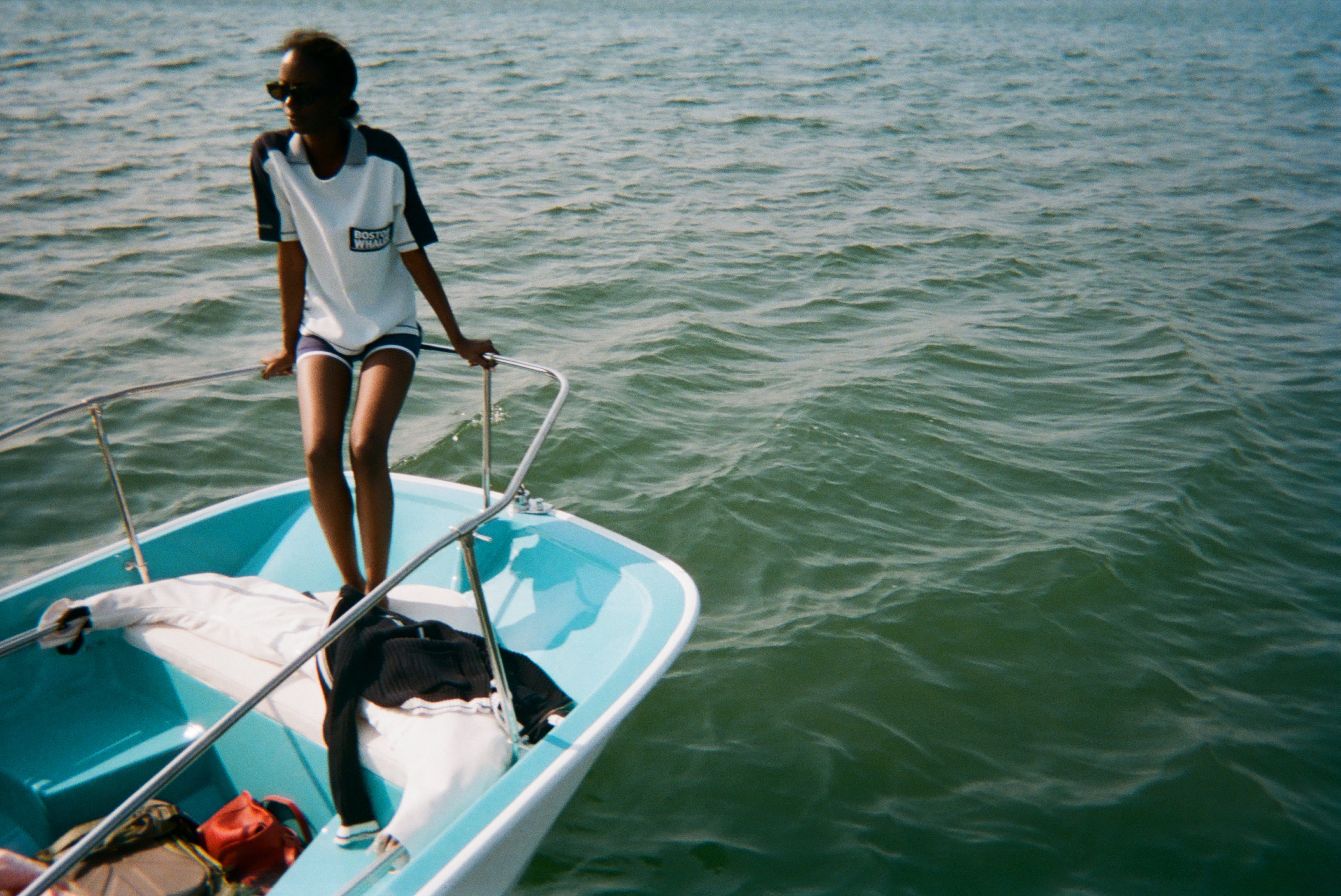 Woman standing at the edge of a boat wearing a light blue cotton mesh shirt with navy shoulder panels and a 'Boston Whaler' logo on the chest, paired with matching navy shorts with white trim. 