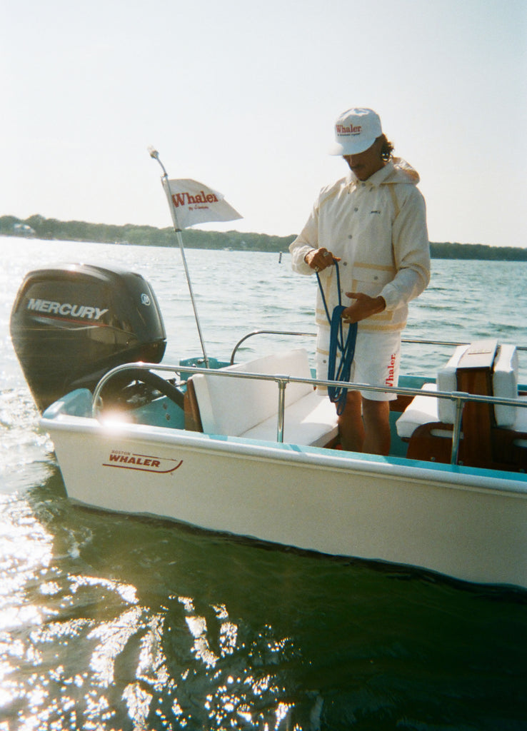 A man standing on a boat in a rain jacket and matching shorts with tan trim. He wears a white cap with red 'Whaler' text.