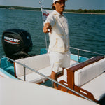 Man standing on a boat in a cream jacket and matching shorts with tan trim, pointing off into the distance. He wears a white cap with red 'Whaler' text.