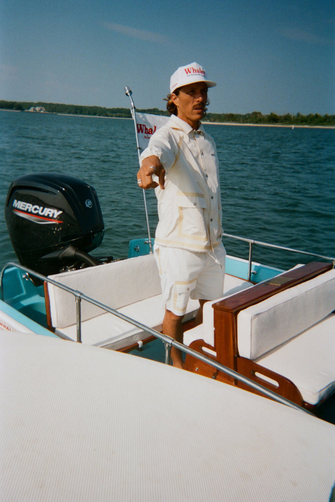 Man standing on a boat in a cream jacket and matching shorts with tan trim, pointing off into the distance. He wears a white cap with red 'Whaler' text.