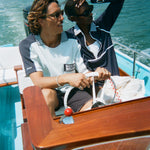 Man and woman riding in a boat. The man wears a light blue cotton mesh shirt with navy shoulder panels and a 'Boston Whaler' logo on the chest. The woman sits beside him in a navy zip-up jacket with white piping and a matching striped collar. A white 'Whaler' flag waves behind them.