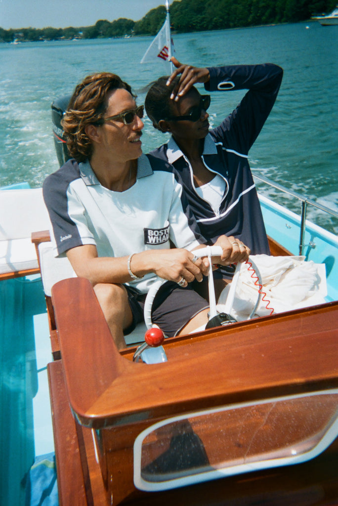Man and woman riding in a boat. The man wears a light blue cotton mesh shirt with navy shoulder panels and a 'Boston Whaler' logo on the chest. The woman sits beside him in a navy zip-up jacket with white piping and a matching striped collar. A white 'Whaler' flag waves behind them.