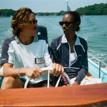 Man and woman riding in a boat. The man wears a light blue cotton mesh shirt with navy shoulder panels and a 'Boston Whaler' logo on the chest. The woman sits beside him in a navy zip-up jacket with white piping and a matching striped collar.