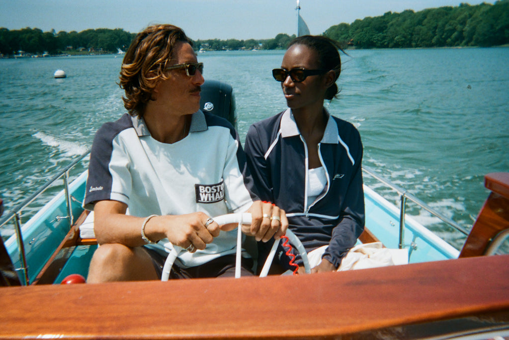 Man and woman riding in a boat. The man wears a light blue cotton mesh shirt with navy shoulder panels and a 'Boston Whaler' logo on the chest. The woman sits beside him in a navy zip-up jacket with white piping and a matching striped collar.