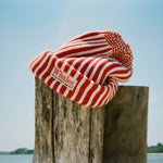 Red and white striped knit beanie with a folded brim and 'Whaler The Unsinkable Legend' patch on the front, draped over a wooden post at the beach.