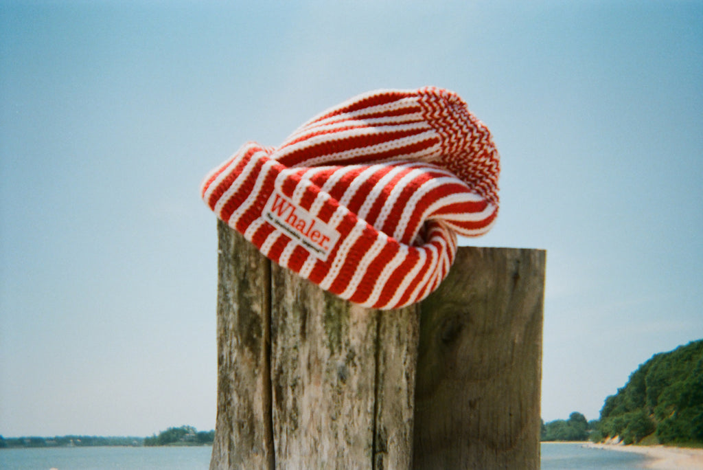 Red and white striped knit beanie with a folded brim and 'Whaler The Unsinkable Legend' patch on the front, draped over a wooden post at the beach.