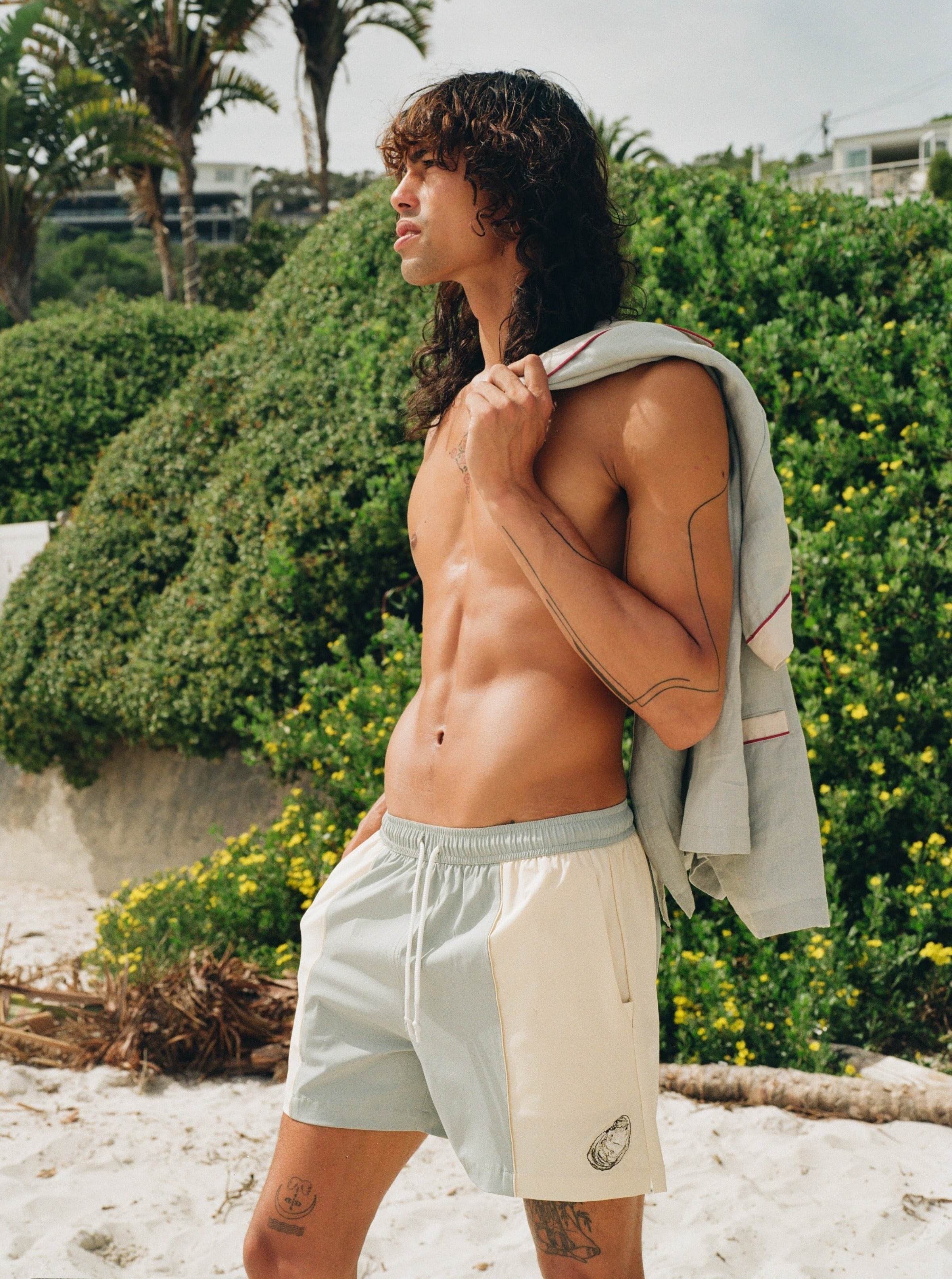 shirtless man wearing Tombolo oyster shirts looks off into distance, holding the linen oyster shirt over his left shoulder