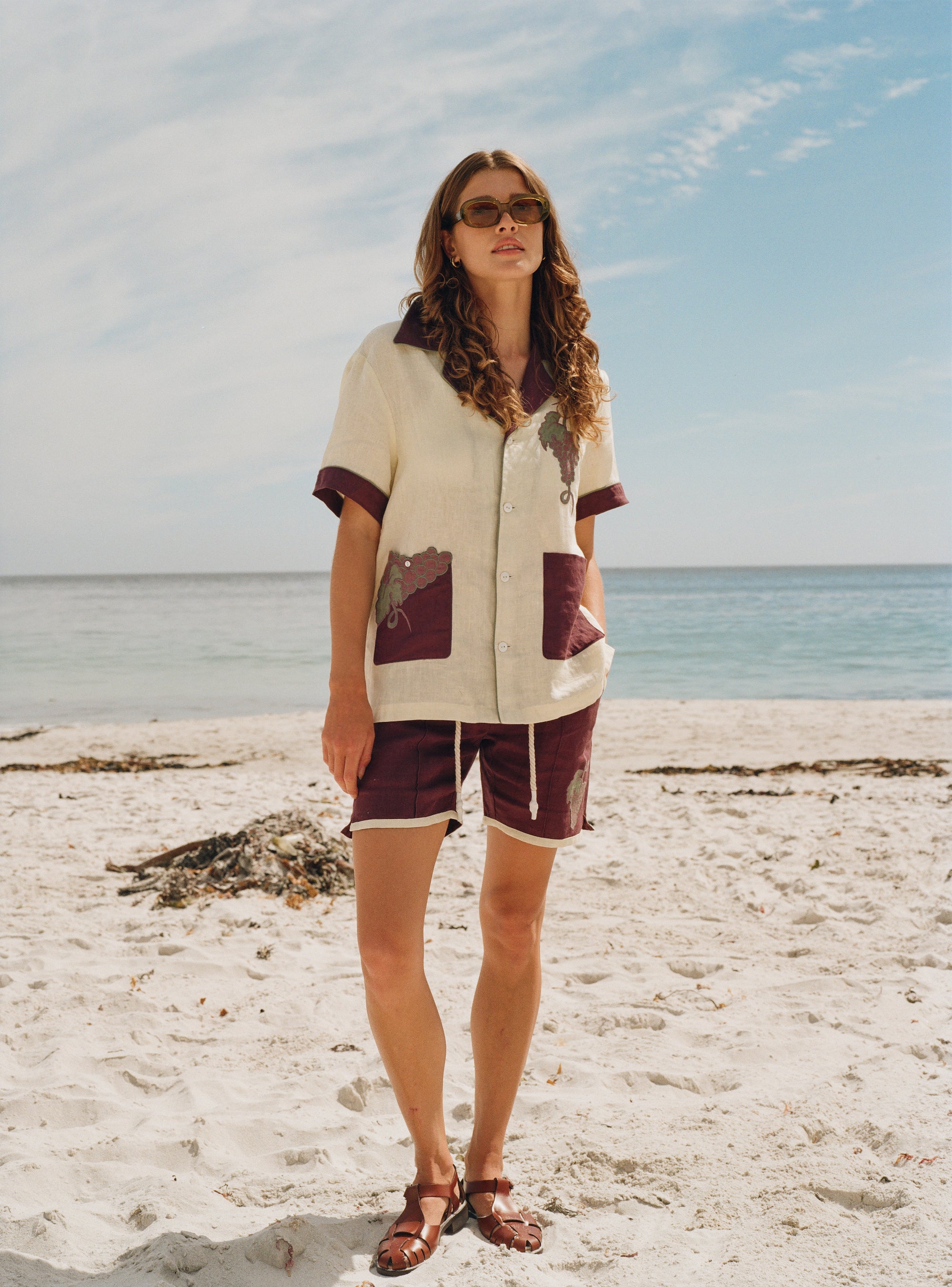 Female model standing on beach wearing full 'Tombarolo' cabana set with sunglasses on and brown leather sandals