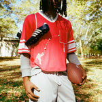 A person wearing a red mesh football-style crop top with cowrie shells forming a football graphic, layered over a gray long-sleeve shirt and paired with gray athletic shorts, holding a football with cleats draped over one shoulder.
