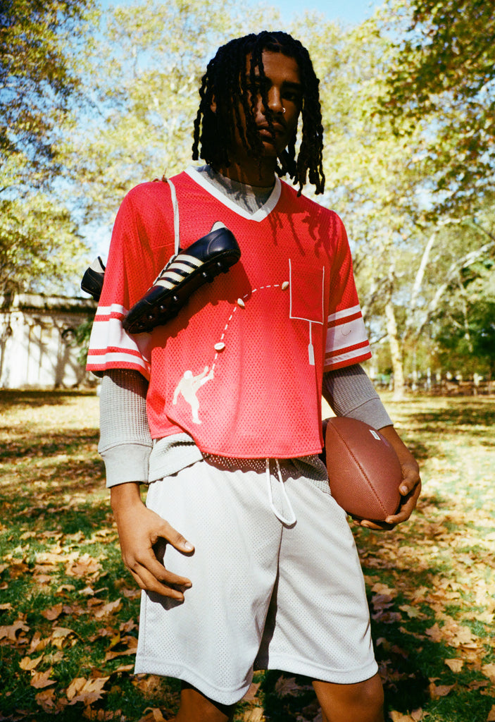 A person wearing a red mesh football-style crop top with cowrie shells forming a football graphic, layered over a gray long-sleeve shirt and paired with gray athletic shorts, holding a football with cleats draped over one shoulder.