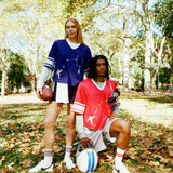 Two people wearing football-style mesh tops with cowrie shells forming a football graphic. One wears a navy blue version with layered long sleeves and shorts, holding a football. The other wears a red version with layered sleeves and shorts, holding a helmet.